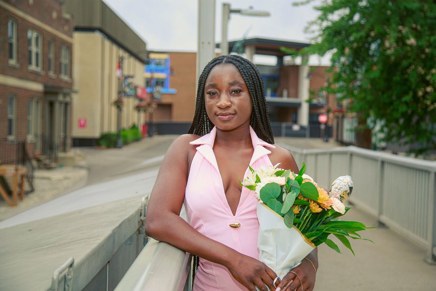 Young woman in pink dress holding yellow and white flowers bouquet, standing on urban bridge walkway.