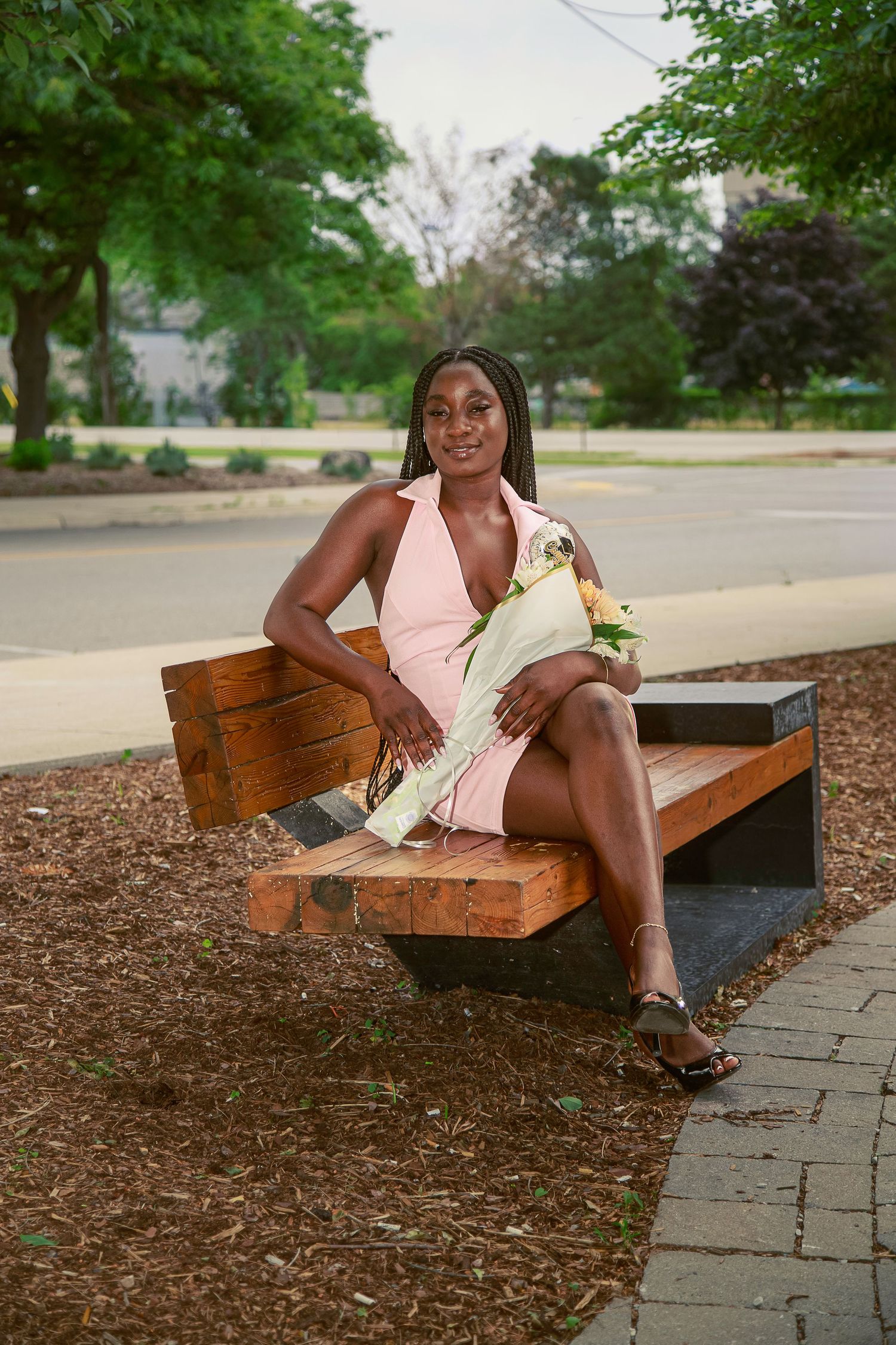 Young woman in pink dress holding flowers, sitting on wooden bench in park, smiling outdoors.