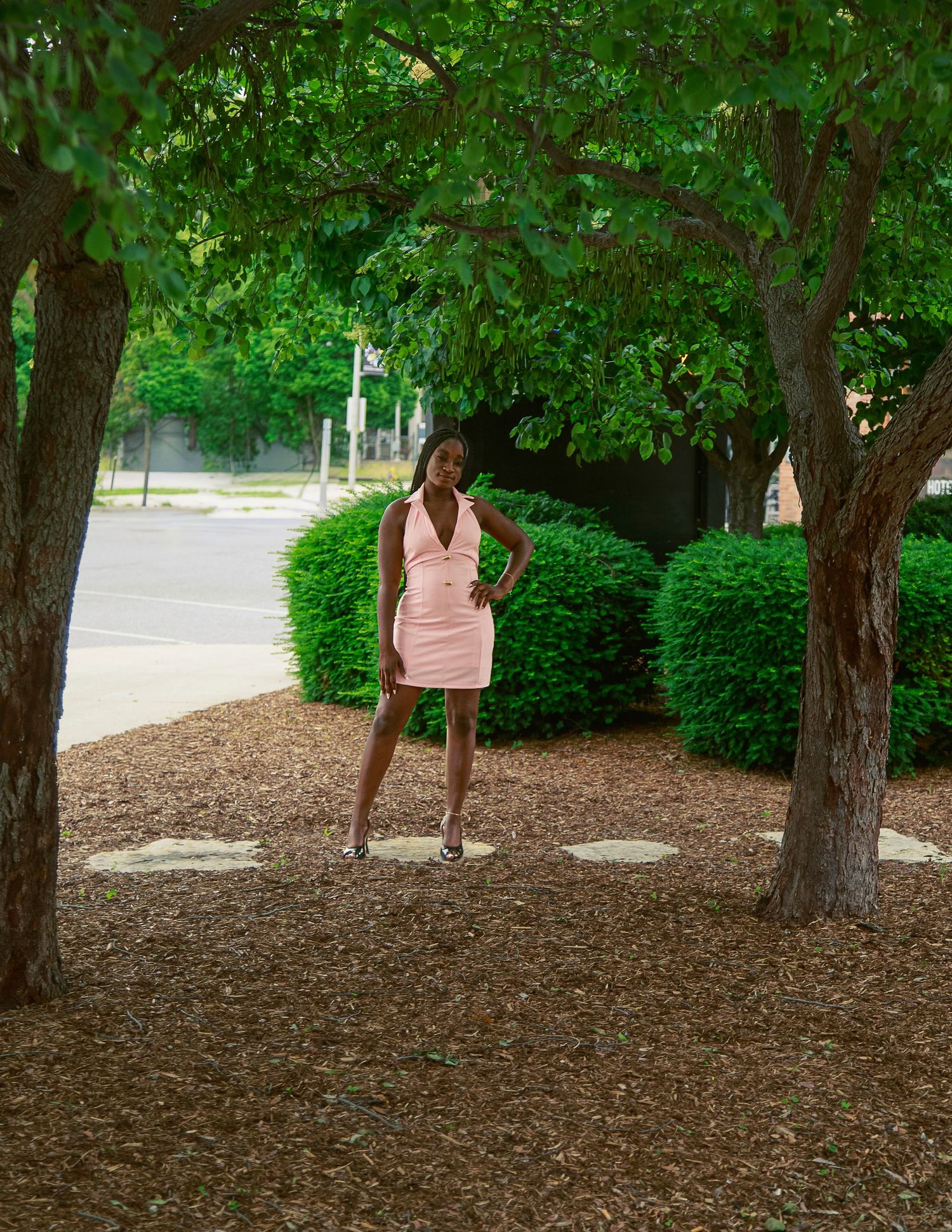 A young woman in a pink mini dress poses confidently between two trees in a landscaped outdoor setting.
