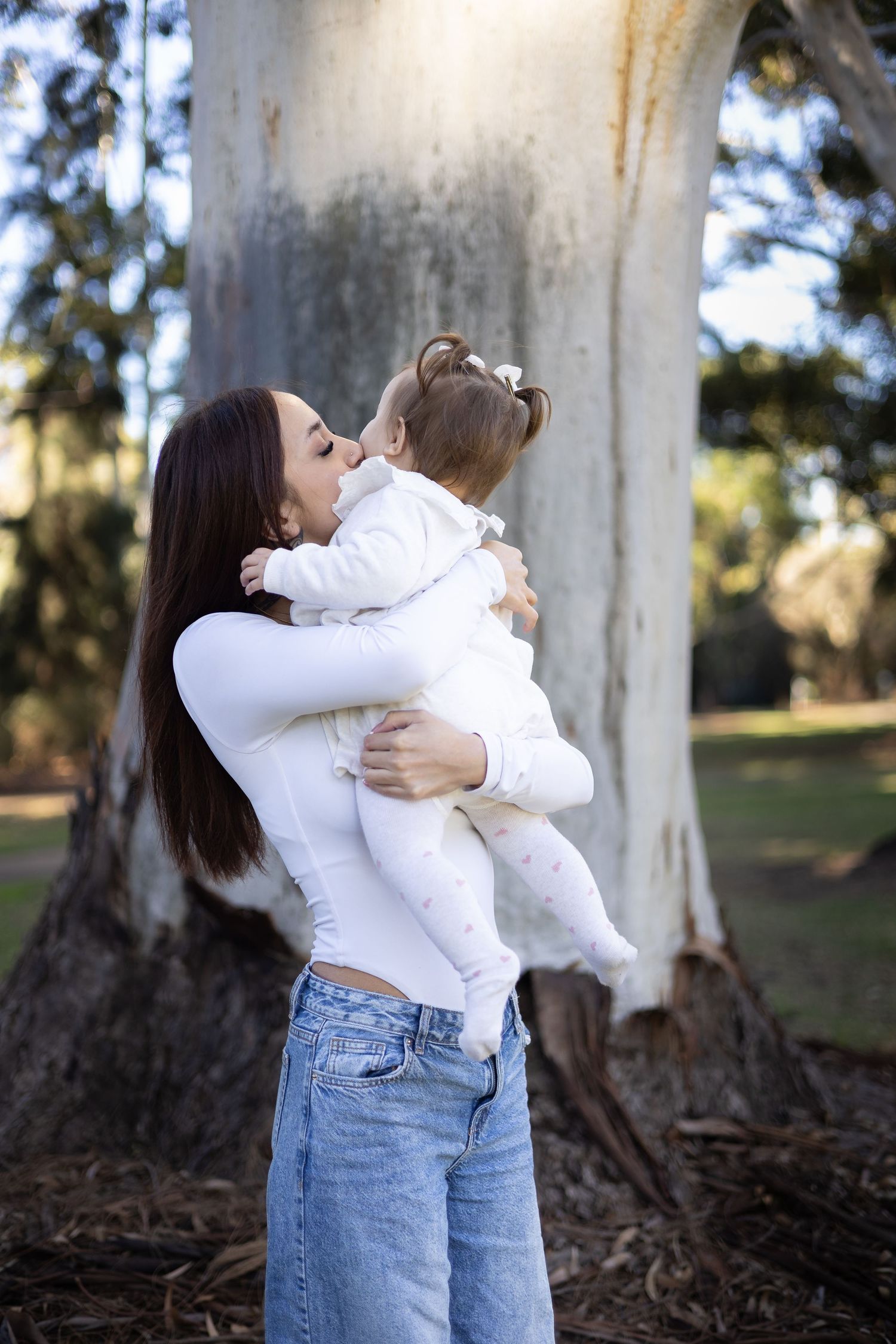 A mother in white top and jeans kisses her baby girl outdoors near a large tree in a sunny park.