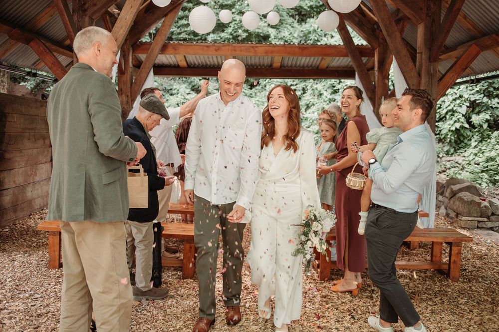 Newlyweds walk down aisle under wooden pavilion as guests celebrate with confetti.
