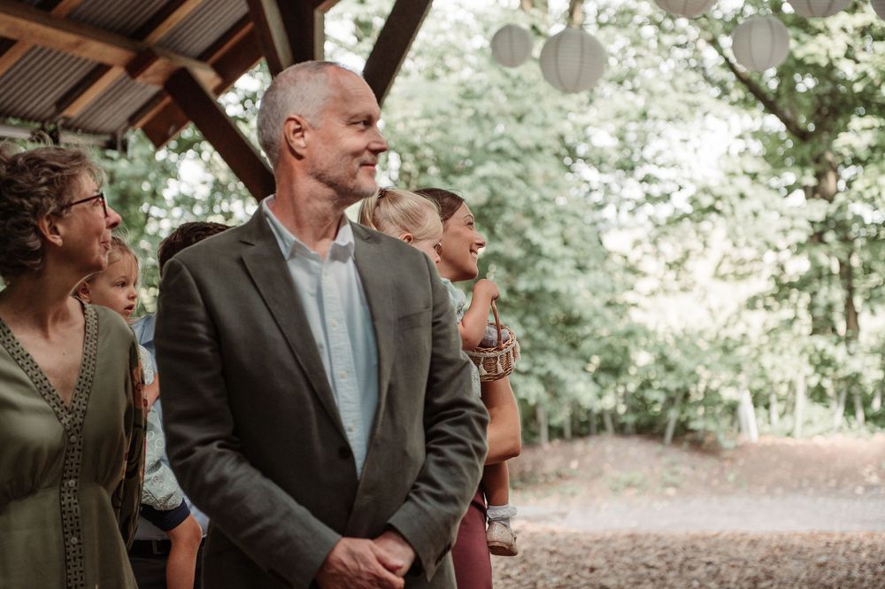 Guests smile during an outdoor wedding ceremony under a wooden pavilion surrounded by lush green trees.