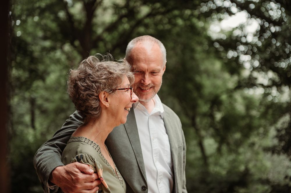 An older couple smiling at each other outdoors, surrounded by lush green trees, dressed in elegant attire.