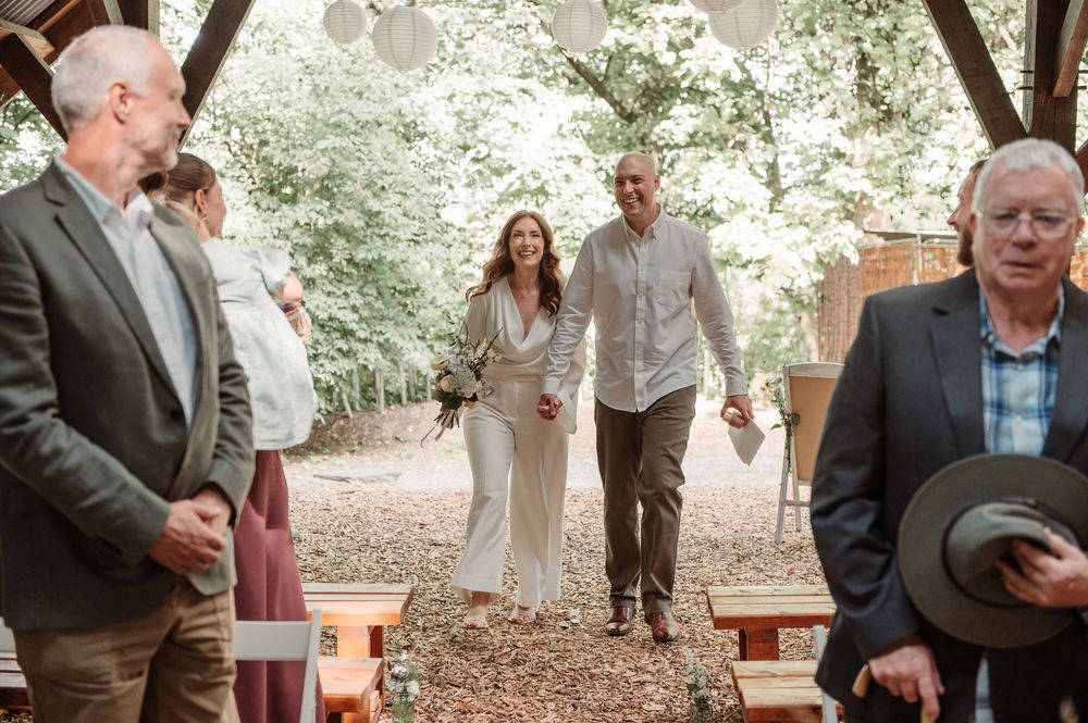 Couple walks hand-in-hand down the aisle at an outdoor woodland wedding ceremony with guests watching.