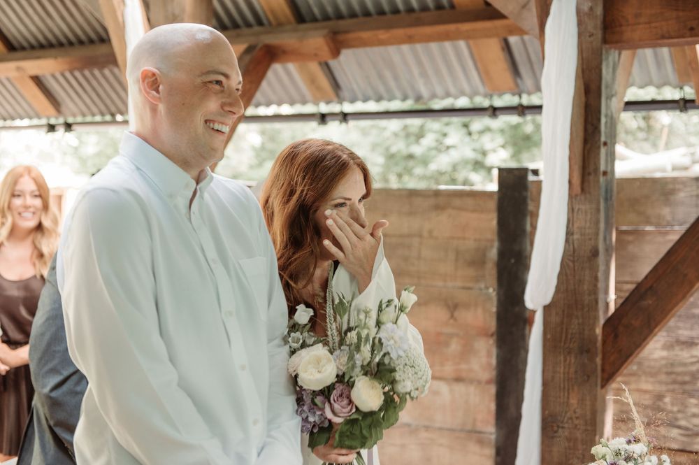 A smiling bald groom and redhead bride holding a floral bouquet share a joyful moment during their rustic barn wedding ceremony.