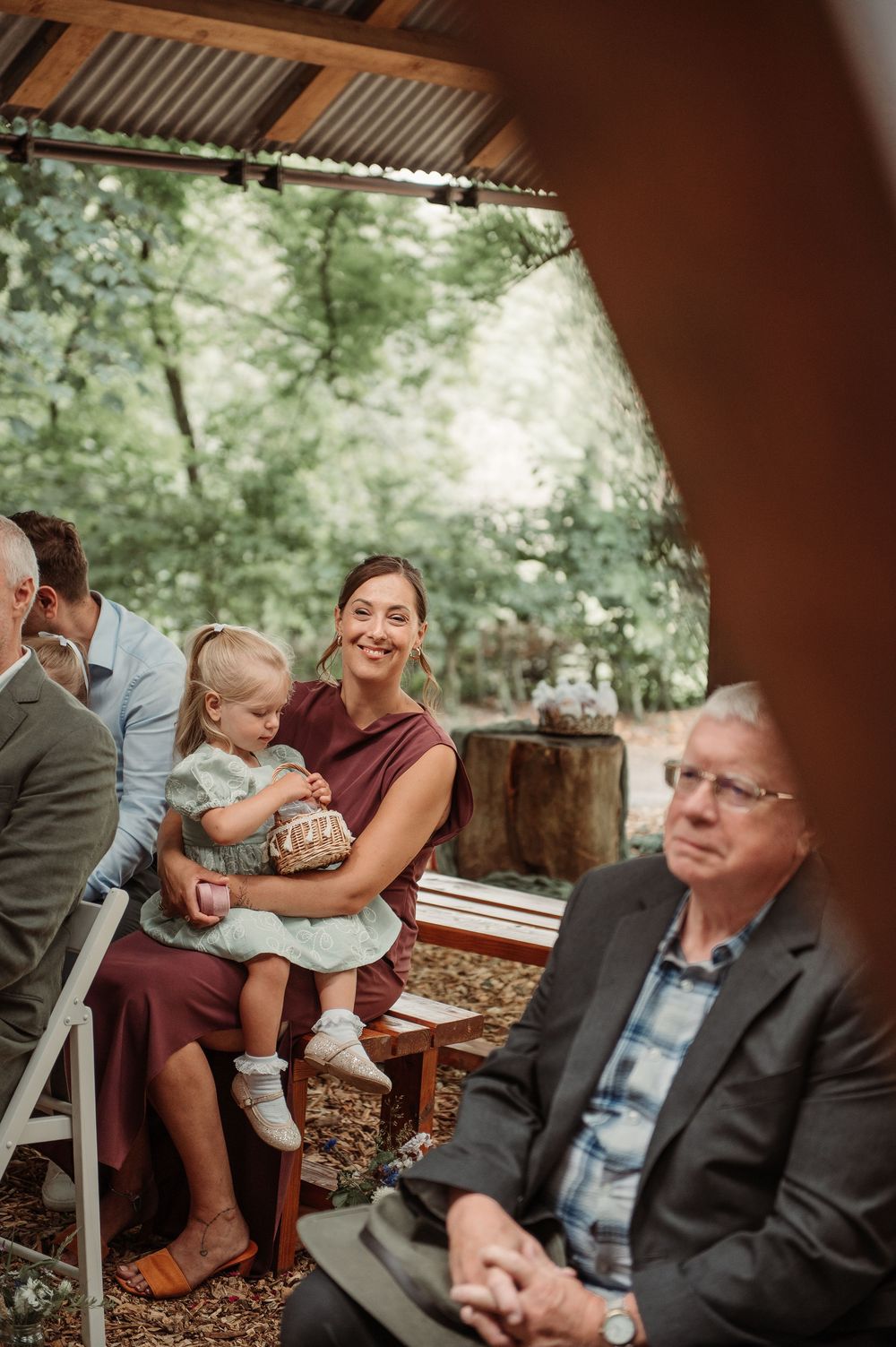 Woman smiling while holding a toddler at an outdoor woodland wedding ceremony with guests seated nearby.