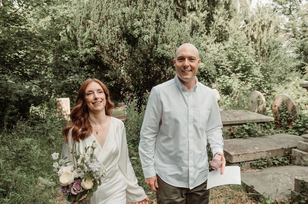 Smiling bride holding bouquet stands with groom in white shirt outdoors among lush greenery.
