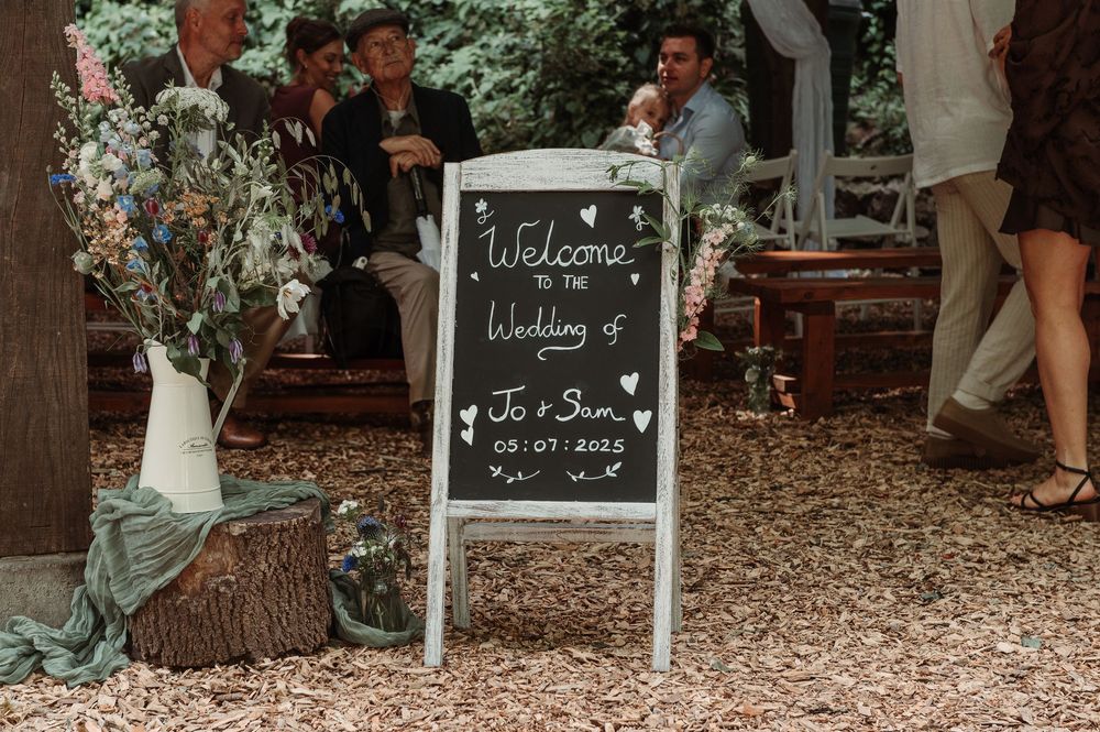 A chalkboard wedding welcome sign reads Welcome to the Wedding of Jo and Ben dated 05.07.2022 at an outdoor venue.