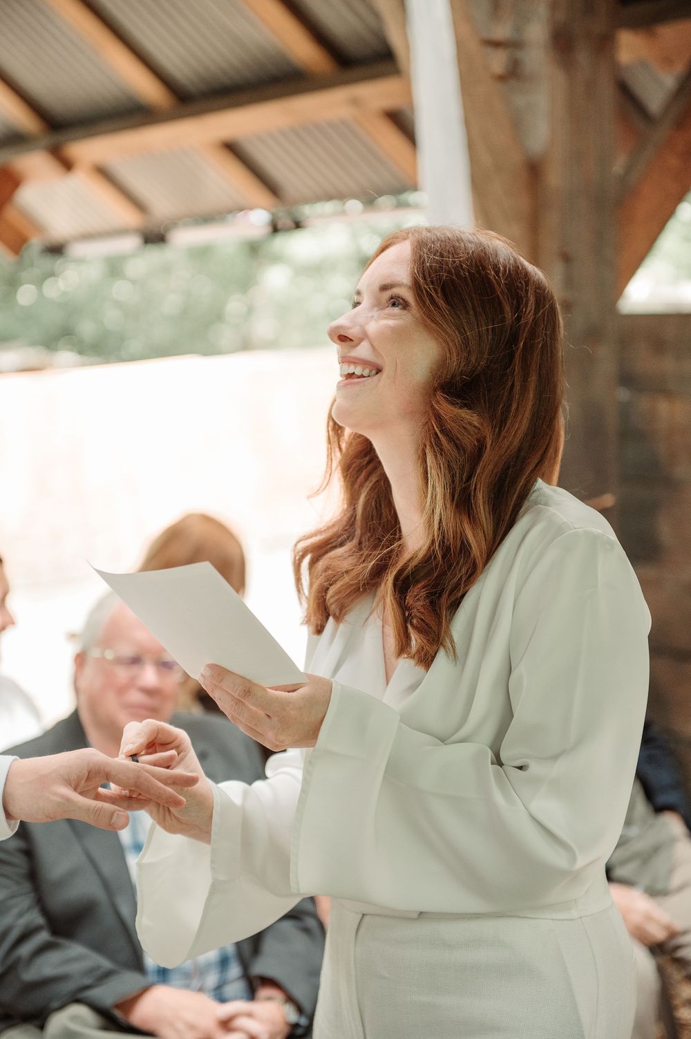 Smiling woman in white blazer holding papers at an outdoor wedding ceremony under a rustic wooden shelter.