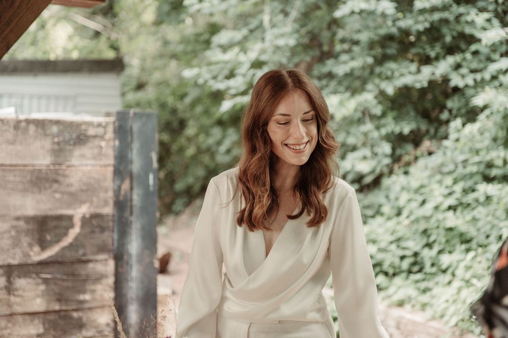 A radiant redhead bride in a white wrap dress smiles softly while standing outdoors surrounded by lush green trees.