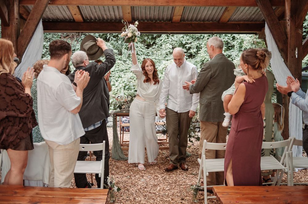 A joyful bride and groom walk back down the aisle as guests applaud at their rustic outdoor wedding ceremony.