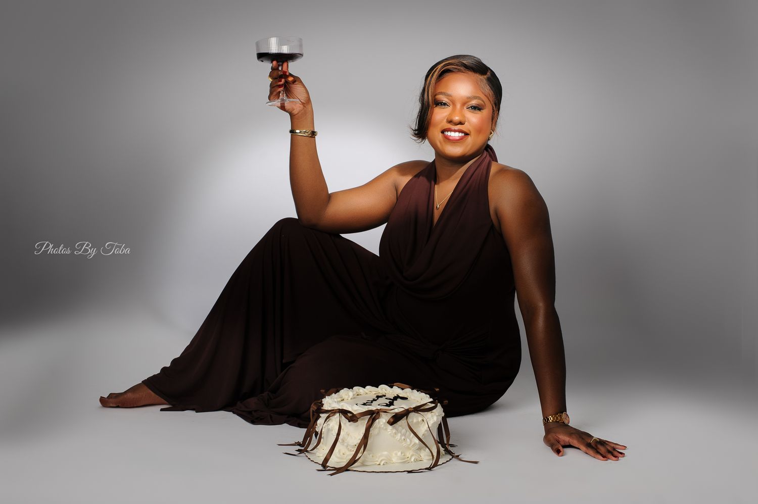 Woman in brown gown sitting on floor, raising wine glass, with white purse, elegant studio portrait photography.