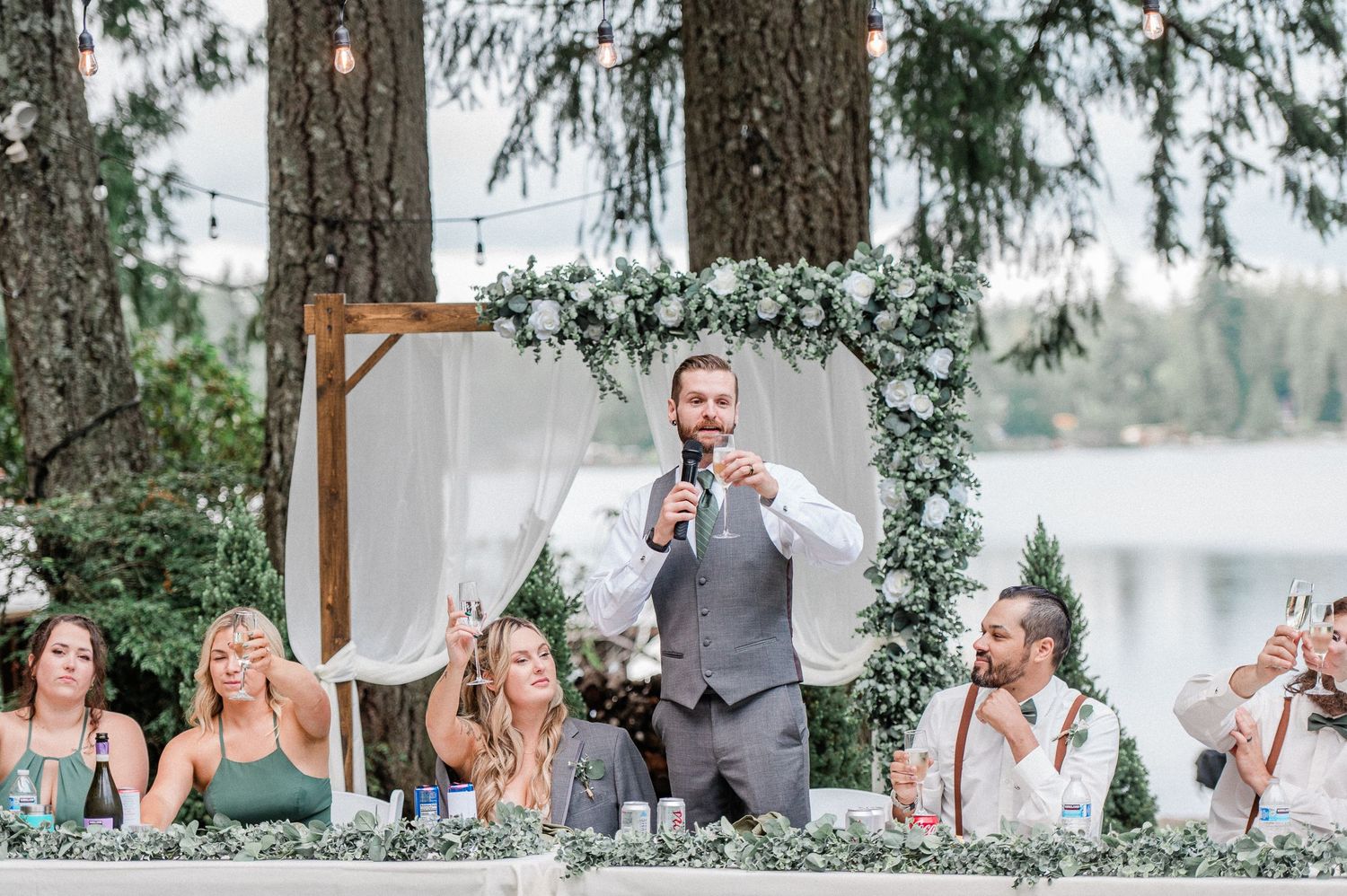 Groom giving a wedding toast speech outdoors under a floral arch with guests holding champagne glasses.