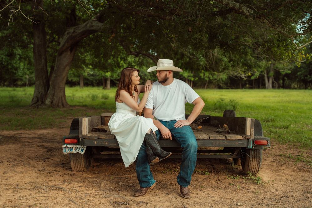 A couple in western attire sits together on a rustic wooden trailer under shady green trees.