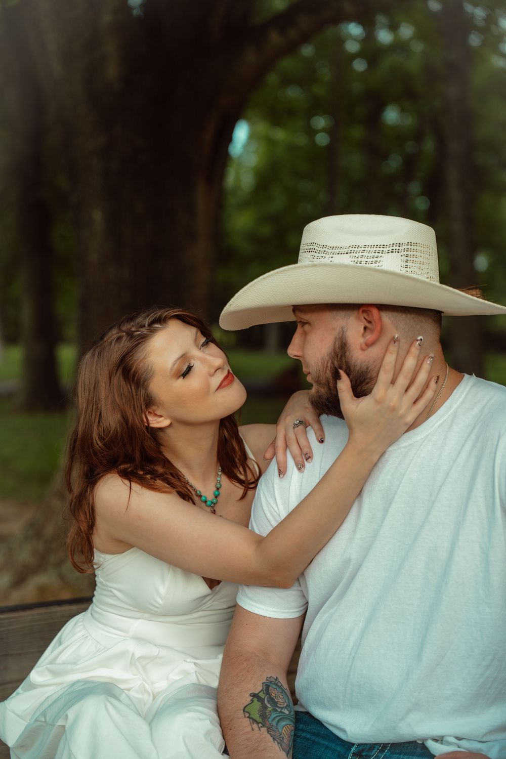 Couple sharing romantic moment in wooded park, woman in white dress touching bearded mans face wearing cowboy hat.