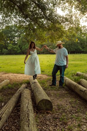 Couple holding hands walking on rustic log bridge in lush green park, woman in white dress, man in cowboy hat.