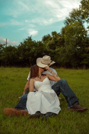 Couple kissing in green field, woman in white dress seated between mans legs, both wearing cowboy boots.
