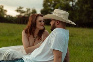 Redhead woman in white dress smiling at bearded man in cowboy hat while sitting together in green field.