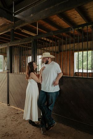 Couple sharing tender moment inside rustic wooden horse stable, woman in white dress touching mans chin.
