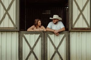 Smiling couple leaning on stable door, woman in white dress and man in cowboy hat in rustic barn setting.
