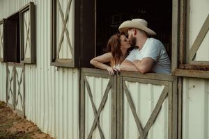 Couple sharing a kiss while leaning over rustic stable door, man in cowboy hat, woman in white dress.