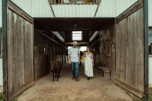 Couple walking miniature horse through rustic barn aisle, woman in white dress and man in cowboy hat.