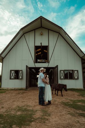 Couple embracing in front of white barn with miniature horse nearby under dramatic blue cloudy sky.