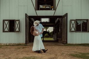 Couple embracing in front of a rustic barn, man in cowboy hat and woman in white dress.