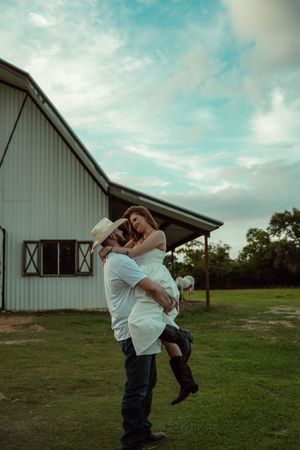 Man in cowboy hat lifting woman in white dress outside a white barn under cloudy sky.