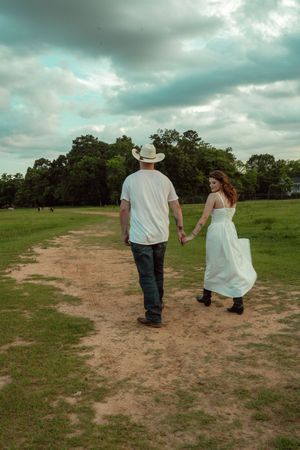 Couple holding hands walking on a dirt path through a green field under cloudy skies.