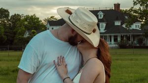 Couple sharing a kiss in a green field with a farmhouse in the background.