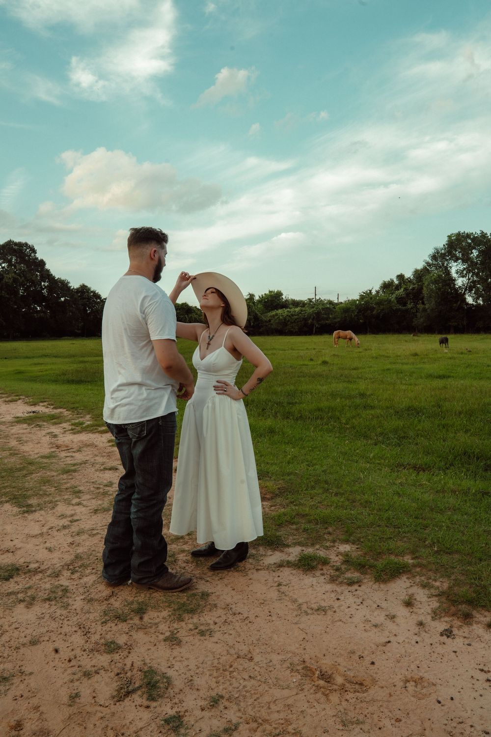 Couple standing in a field, man placing cowboy hat on woman in white dress.