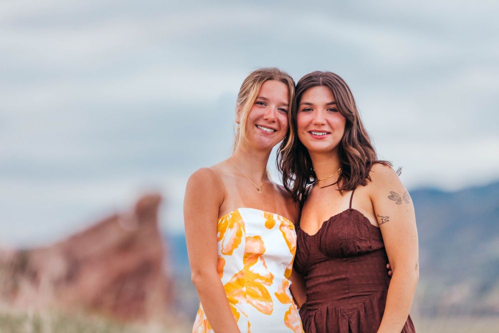 Two smiling women posing together outdoors near red rock formations under a cloudy sky.