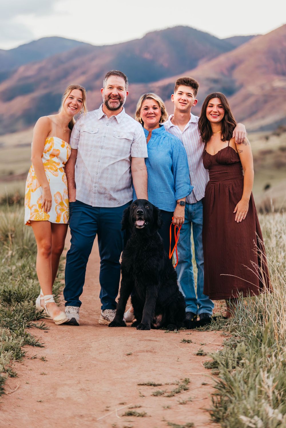 Happy family of five posing with black dog on mountain trail at sunset.