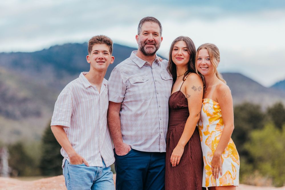 Family of four smiling outdoors with mountain backdrop.