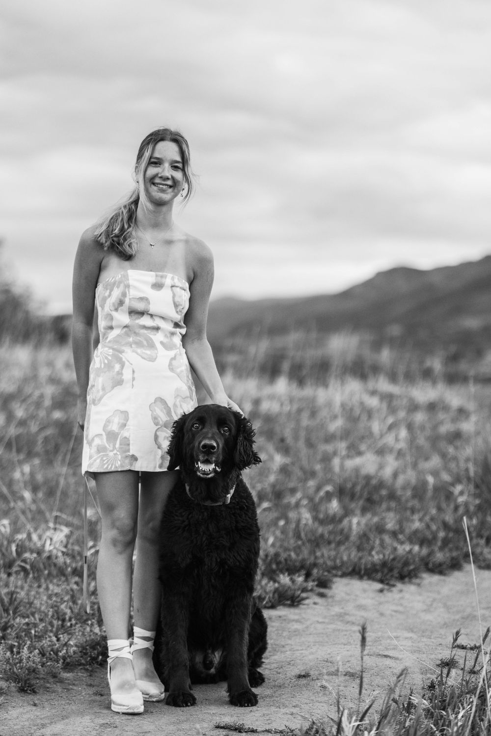 Young woman in floral dress smiling outdoors with her black Cocker Spaniel dog on a rural path.