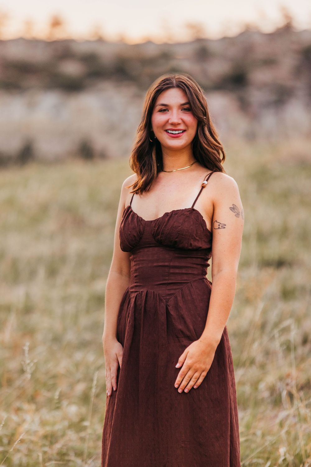 Young woman in brown dress smiling in a field at sunset.