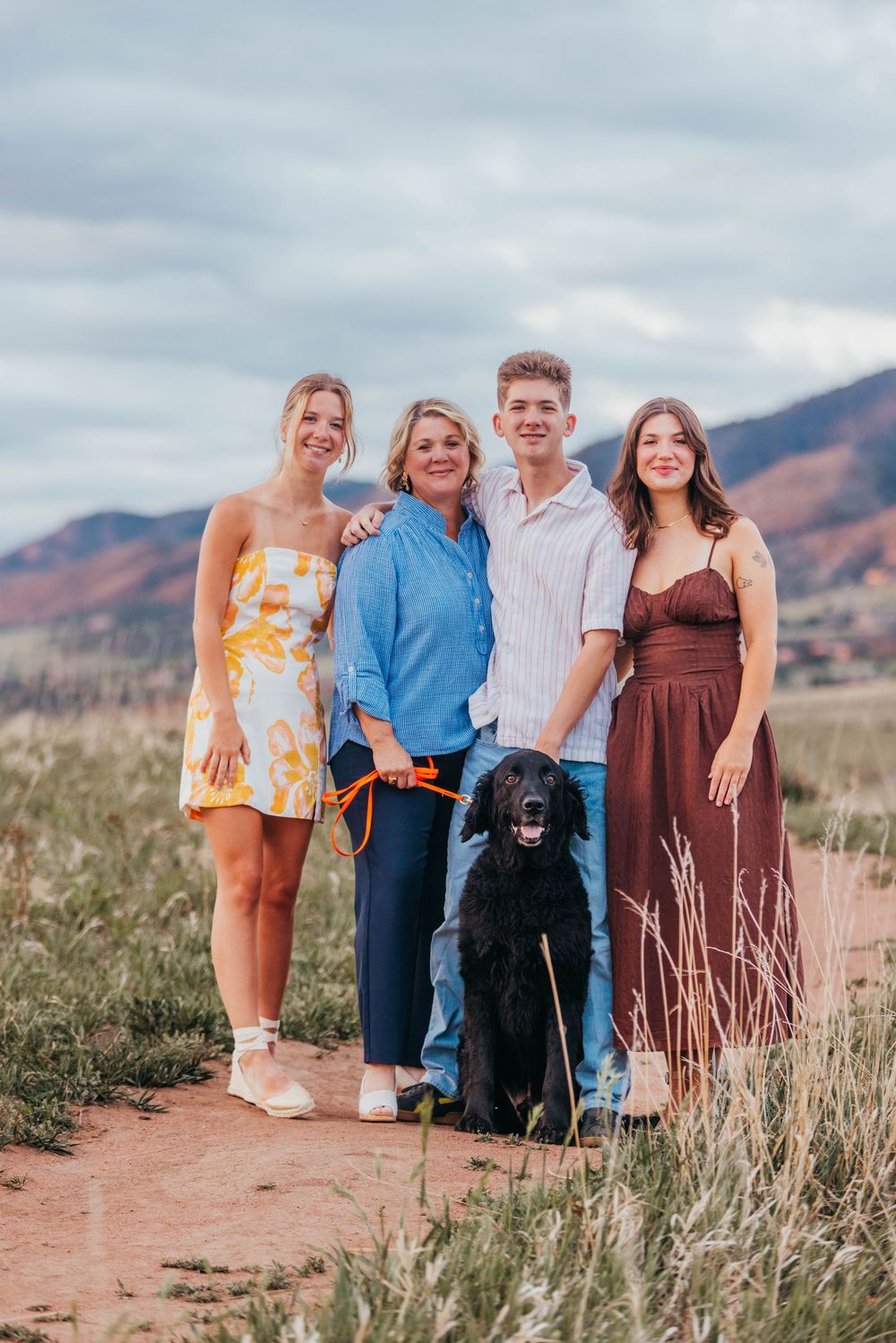 Family of four poses with black dog on mountain trail, wearing summer outfits against scenic Colorado landscape.