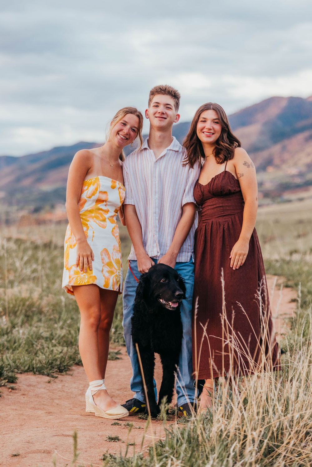 Two young women and a man pose with a black dog in a scenic mountain field under a cloudy sky.