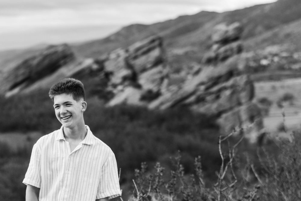 Smiling teenage boy in striped shirt standing in front of rocky mountain landscape in black and white.