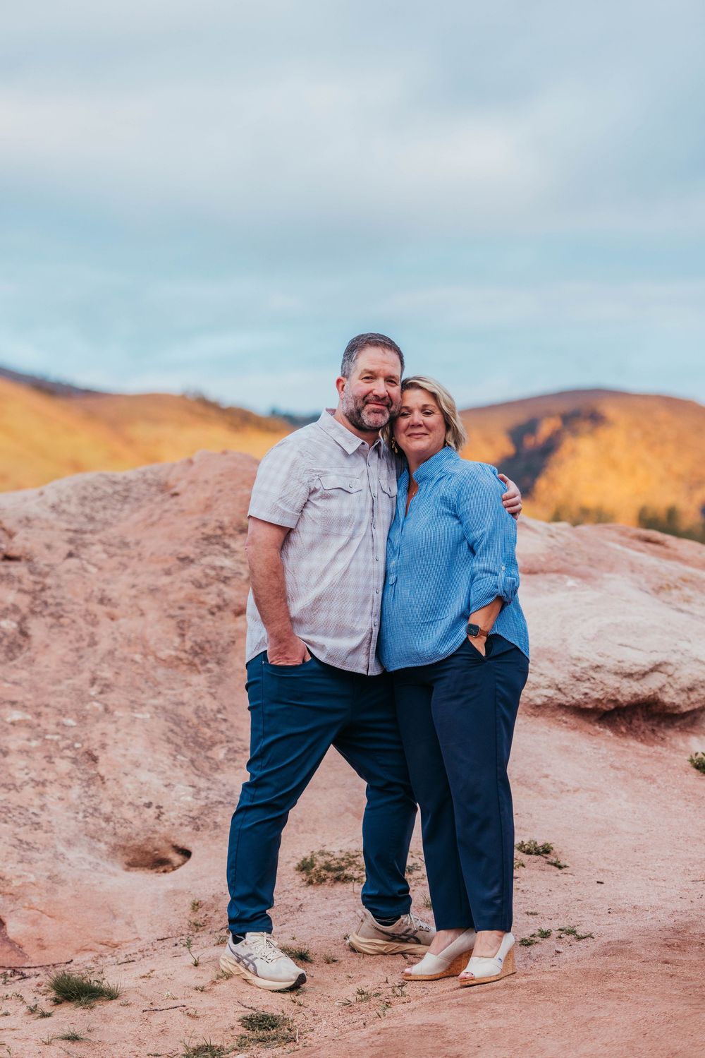Couple embracing and smiling on red rock trail with autumn foliage and hills in background.