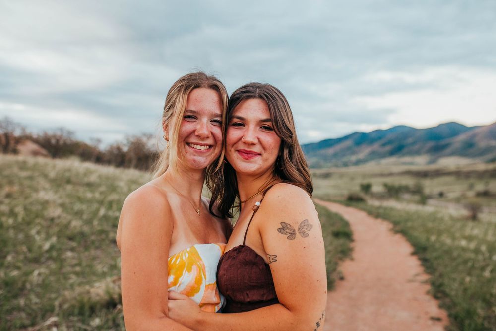 Two smiling women hugging outdoors on a dirt path with green fields and mountains in the background.