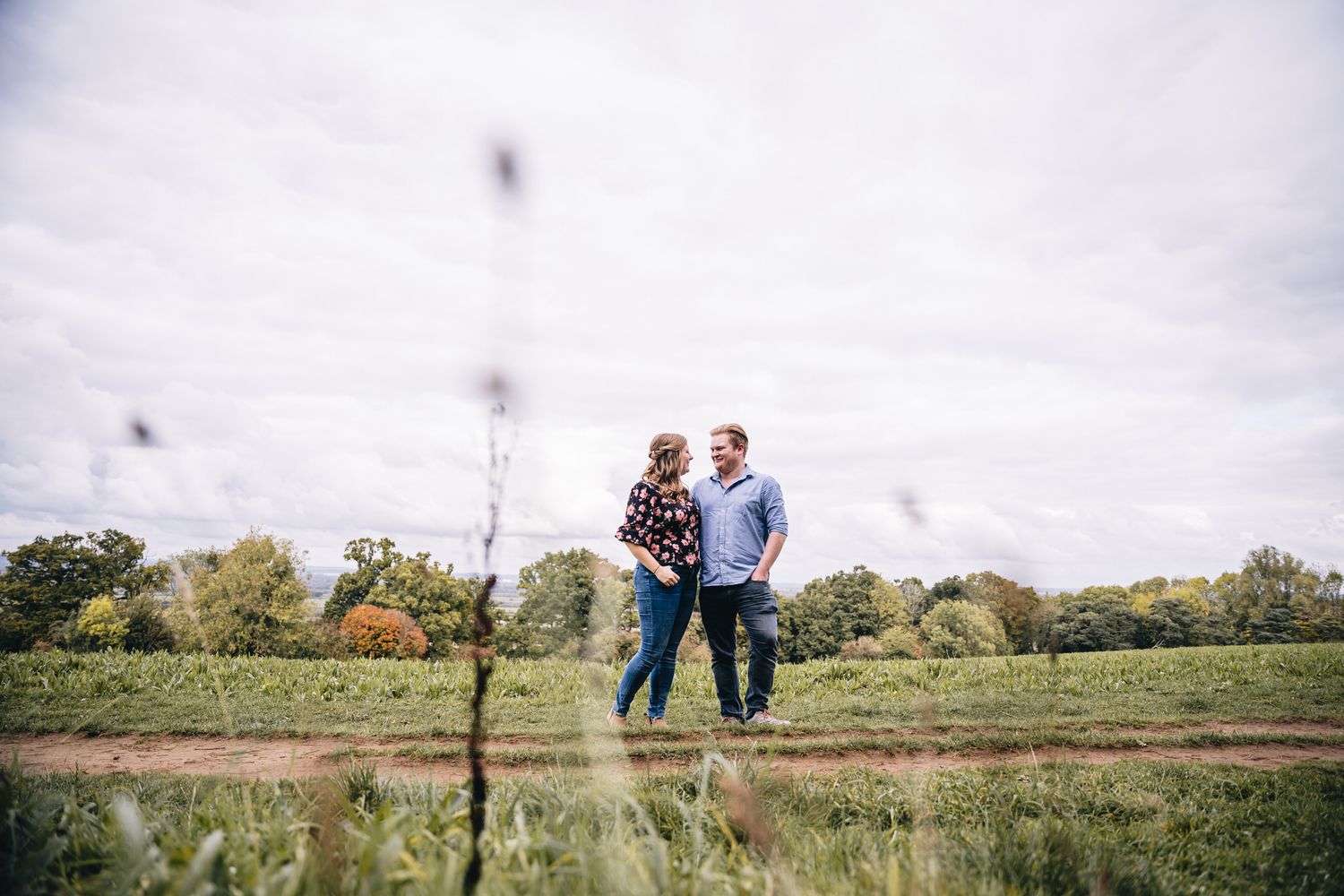 Couple standing together in a lush green field under cloudy skies, sharing a romantic outdoor moment.