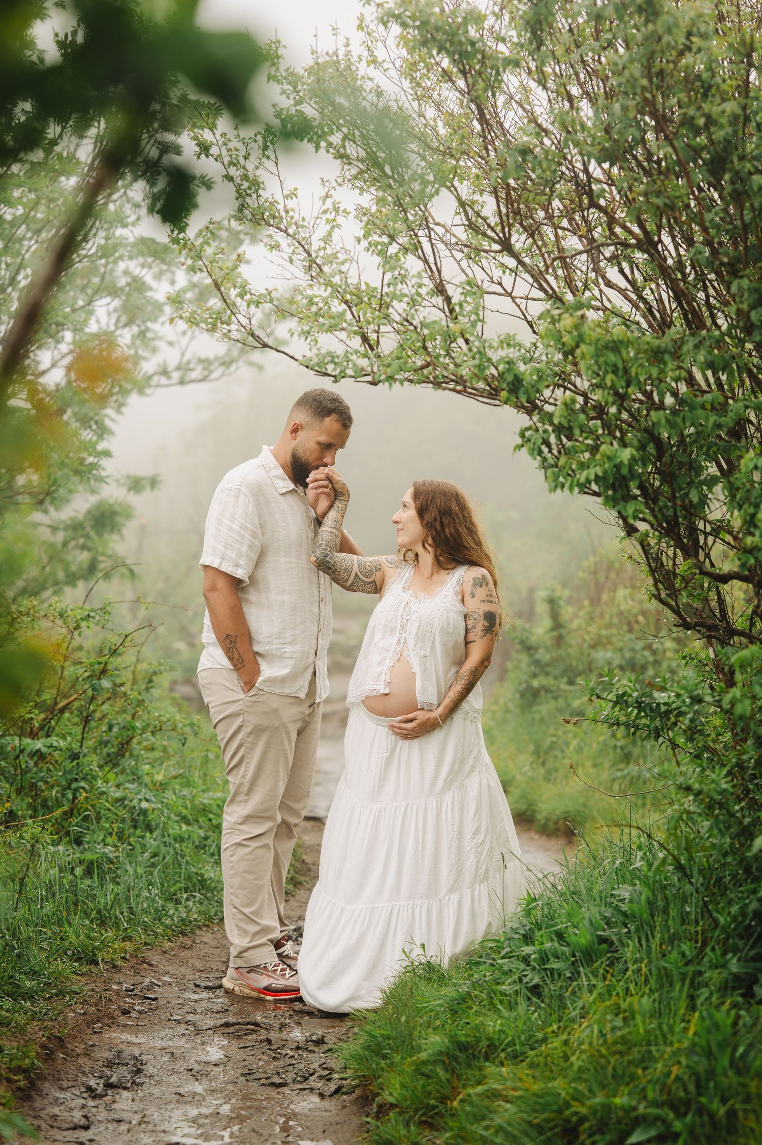 Couple in white outfits on misty forest path, pregnant woman in lace dress holding belly while man tenderly holds her hand.