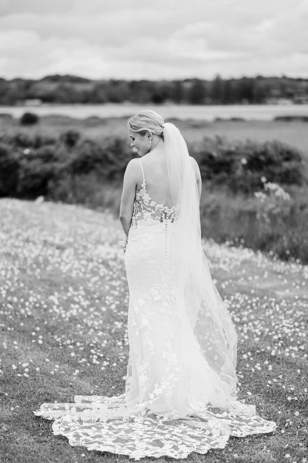 Bride in elegant lace wedding dress with veil standing in a flower field by a lake, black and white photo.