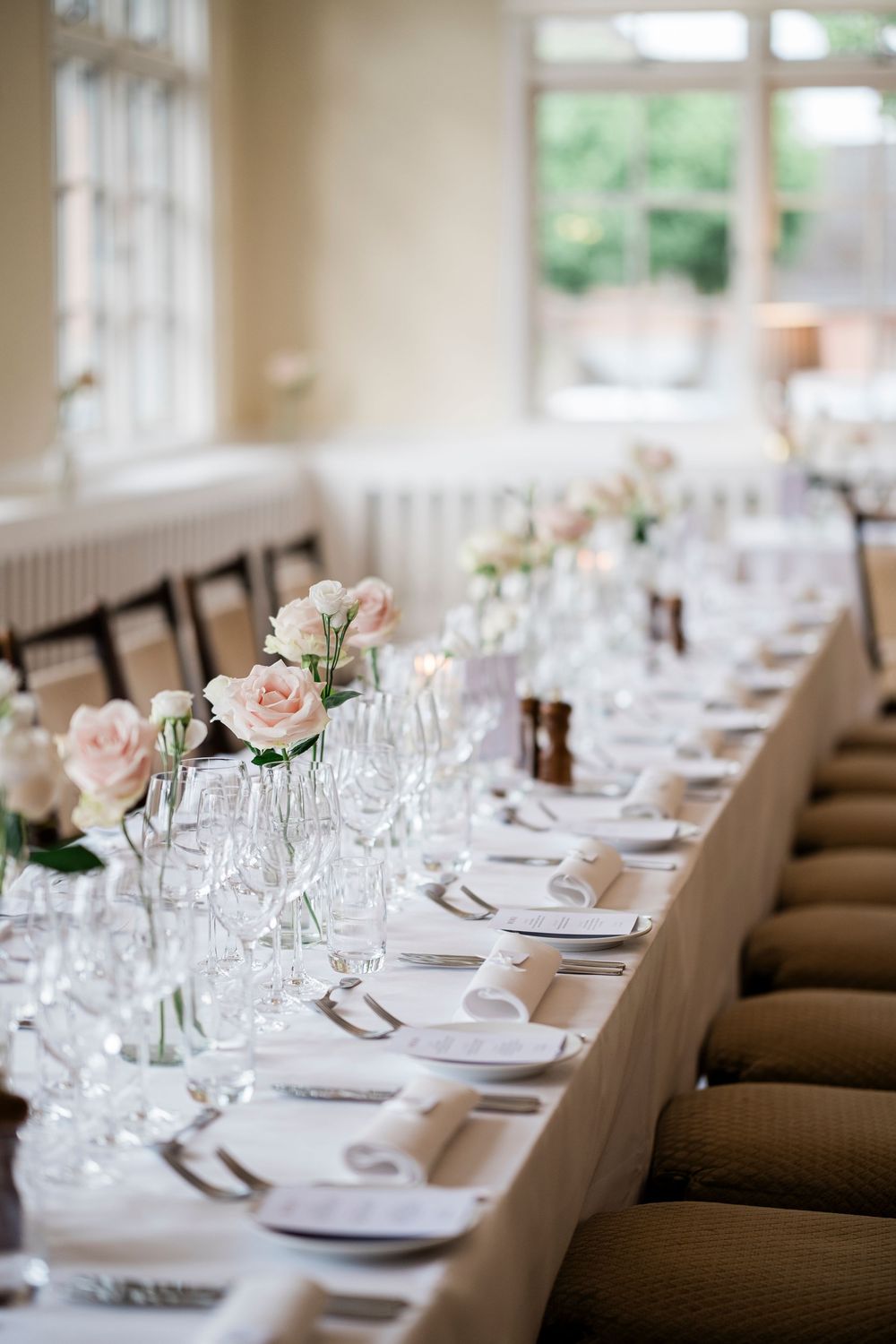 Elegant wedding reception table with pink roses, crystal glasses, white linens, and place settings in a bright venue.