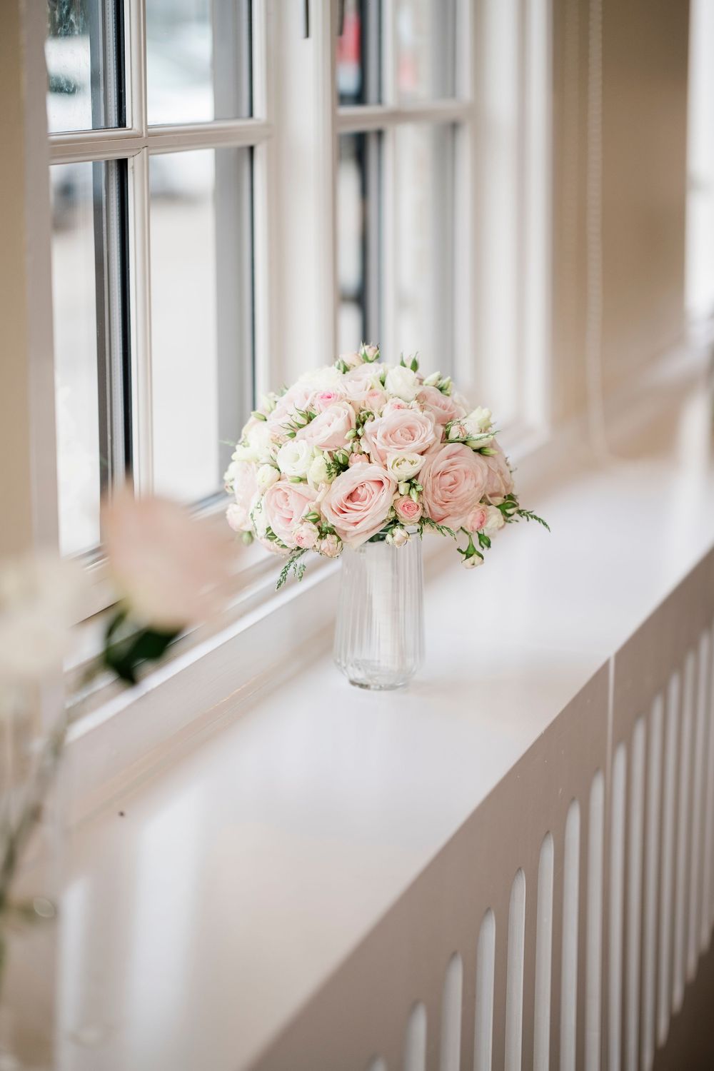 Elegant bridal bouquet of soft pink roses and white flowers in a glass vase on a white window sill.