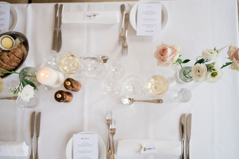 Elegant wedding table setting with white linens, blush roses, candles, wine glasses, and gold cutlery viewed from above.