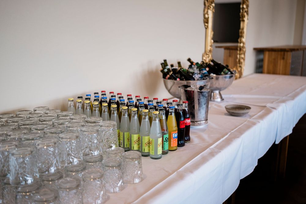 Beverage table with assorted soft drink bottles, empty glasses, and a champagne bucket at a catered event.