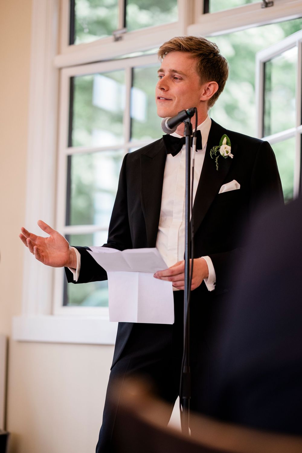 Elegant groom in black tuxedo delivering a heartfelt speech at a microphone during wedding reception.